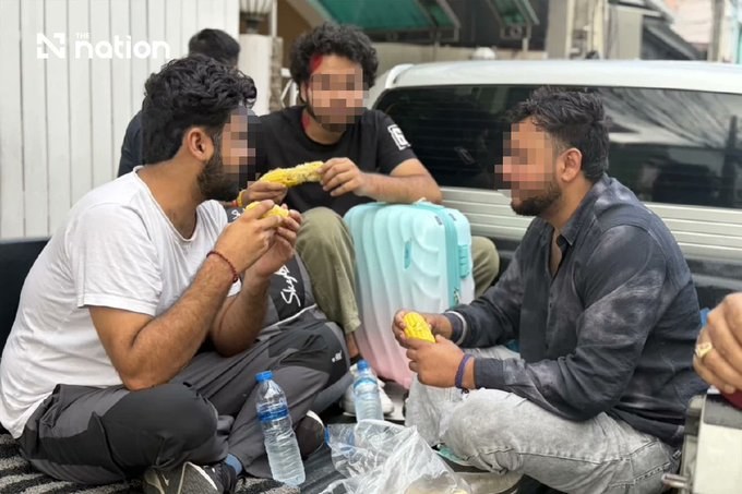 The three Indian tourists rescued in Pattaya Thailand sitting on the ground beside their suitcases and eating corn shortly after police freed them from the Nong Prue townhouse, faces obscured by the news agency