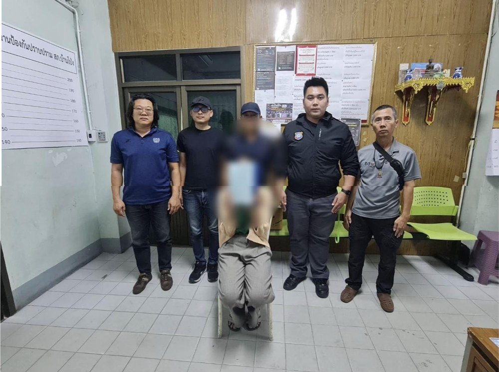 Thai news photograph of police officers standing with a seated suspect at Ban Pong police station in Ratchaburi after an arrest linked to the Pattaya Thailand tourist robbery case