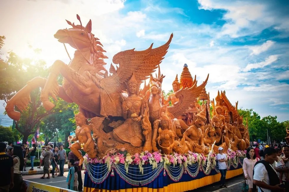 A carved beeswax float topped by a winged horse and celestial figures, garlanded with flowers, waiting on the street among spectators at the Ubon Ratchathani candle procession